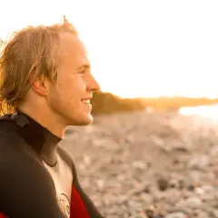 Uśmiechnięty surfer w piance, na planie średnim, patrzy w stronę zachodzącego słońca na kamienistej plaży.