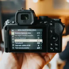 Ręka trzyma bezlusterkowiec pełna klatka, na ekranie menu nagrywania wideo. Ranking aparatów jest w tle.