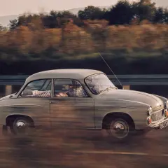 Klasyczny samochód w ruchu, uchwycony techniką panoramowania. W tle rozmyte drzewa i wzgórza.