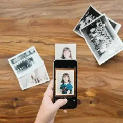Skan ze zdjęcia: dłoń trzyma smartfon, który fotografuje zdjęcie dziewczynki. Wokół leżą stare fotografie.