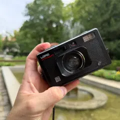 Czarna, analogowa lomografia LomoApparat z obiektywem 21mm. W dłoni, na tle zieleni i fontanny.