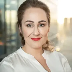 Portret fotografia: uśmiechnięta kobieta z burzą loków, w białej bluzce, z czerwonymi ustami, na tle rozmazanego miasta.