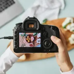 Ręce trzymają aparat cyfrowy, który robi zdjęcie kompozycji z figami i pomidorkami.