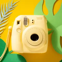 Żółty aparat Fujifilm Instax mini 8, z którego wychodzą zdjęcia, otoczony zielonymi liśćmi monstery i niebieskimi liśćmi paproci na żółtym tle.