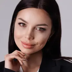Kobieta w czarnej marynarce, z dłonią pod brodą, pokazuje, jak zrobić zdjęcie portretowe. Jej spojrzenie jest ciepłe, a usta lekko uchylone.