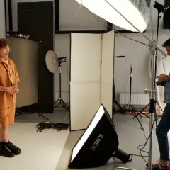 Model w pomarańczowym stroju pozuje, a fotograf ustawia sprzęt. W tle widać lampy i dyfuzory, tworzące idealne światło kontrowe.