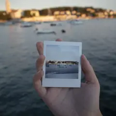 Dłoń trzyma zdjęcie jak z Instax z widokiem na zatokę z łodziami i miasteczkiem w tle.
