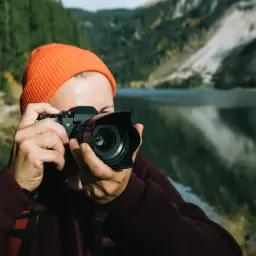 Osoba w pomarańczowej czapce robi zdjęcie aparatem, szukając idealnej lustrzanki na początek. W tle jezioro i góry.