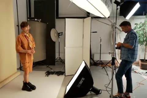 Model w pomarańczowym stroju pozuje, a fotograf ustawia sprzęt. W tle widać lampy i dyfuzory, tworzące idealne światło kontrowe.