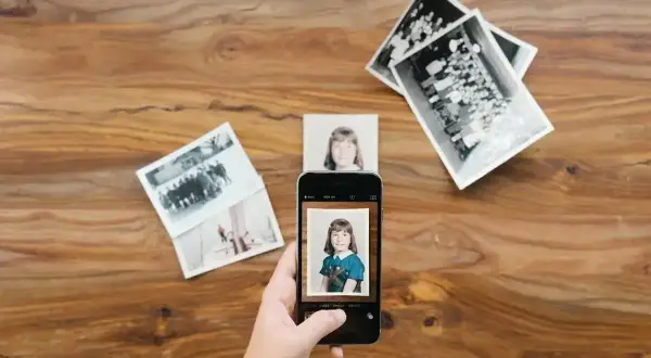 Skan ze zdjęcia: dłoń trzyma smartfon, który fotografuje zdjęcie dziewczynki. Wokół leżą stare fotografie.
