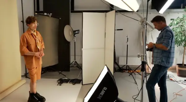 Model w pomarańczowym stroju pozuje, a fotograf ustawia sprzęt. W tle widać lampy i dyfuzory, tworzące idealne światło kontrowe.