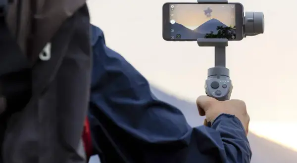 Osoba trzyma smartfon na gimbala, nagrywając piękny krajobraz z górami i zachodzącym słońcem.