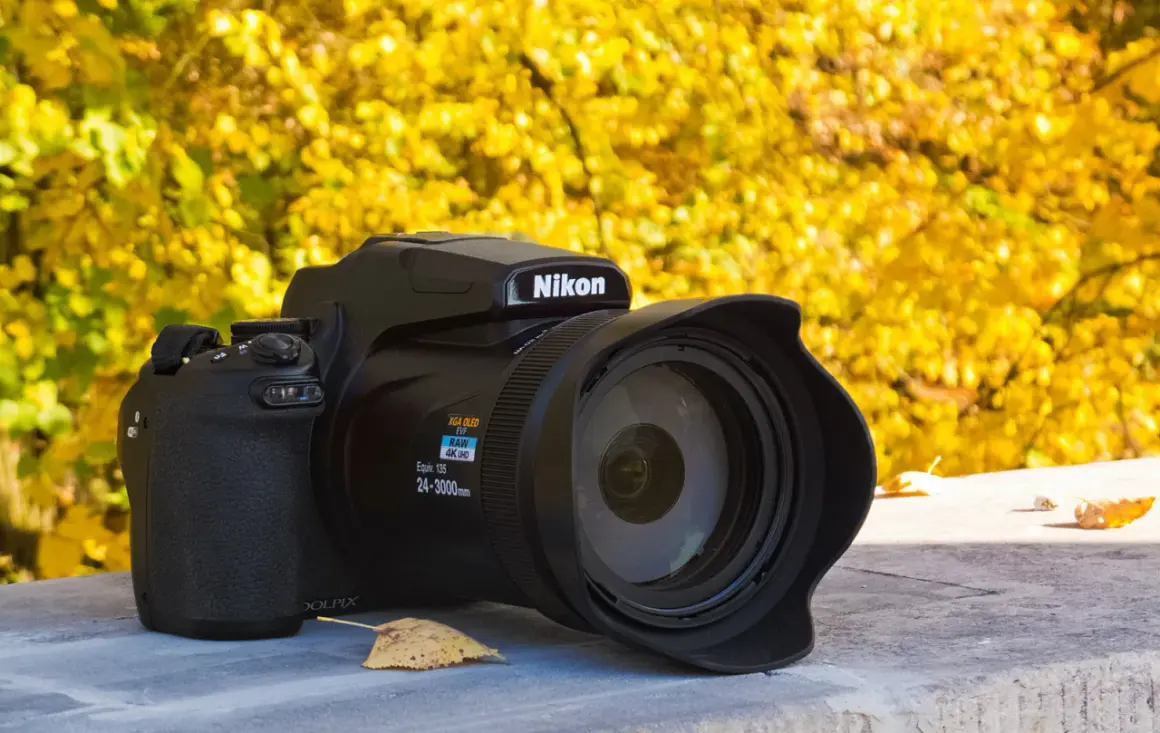 Czarny aparat Nikon P1000 z obiektywem na tle jesiennych, żółtych liści.