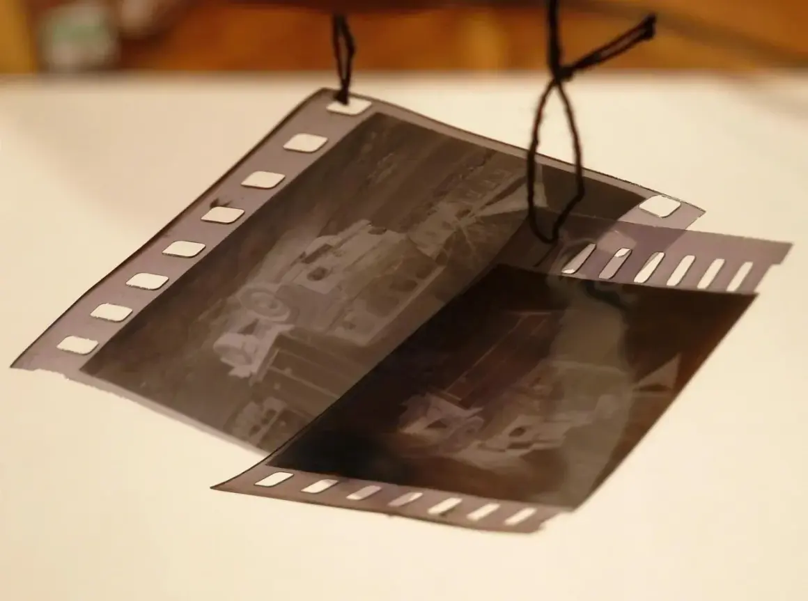Wywoływanie zdjęć z kliszy. Dwie taśmy filmowe z widocznymi klatkami, wiszące na czarnych nitkach.