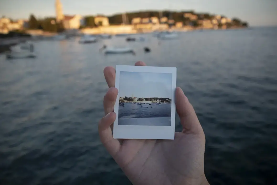 Dłoń trzyma zdjęcie jak z Instax z widokiem na zatokę z łodziami i miasteczkiem w tle.