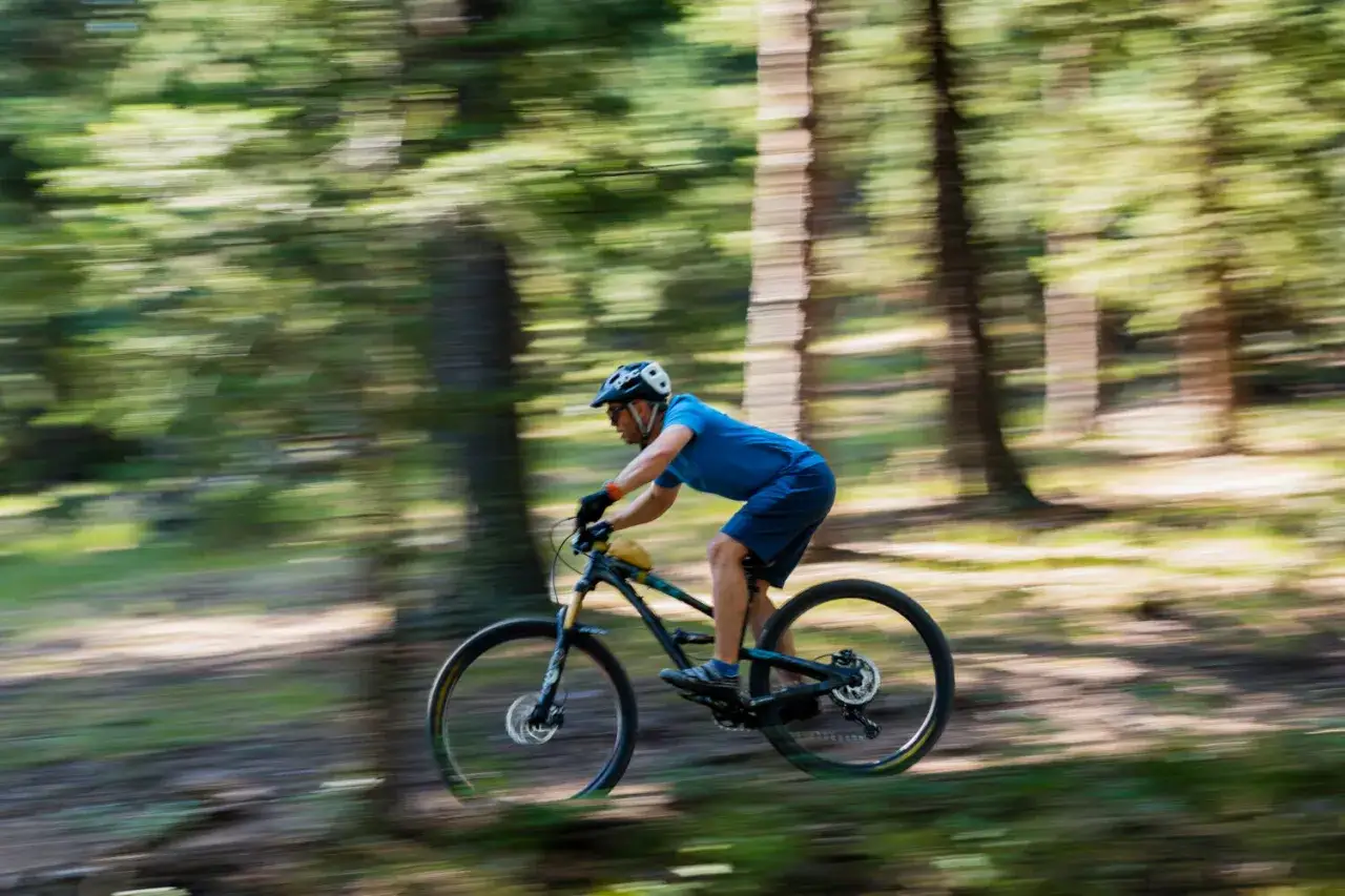 Rowerzysta w niebieskiej koszulce i kasku pędzi przez las. Efekt panoramowania podkreśla dynamikę jazdy.