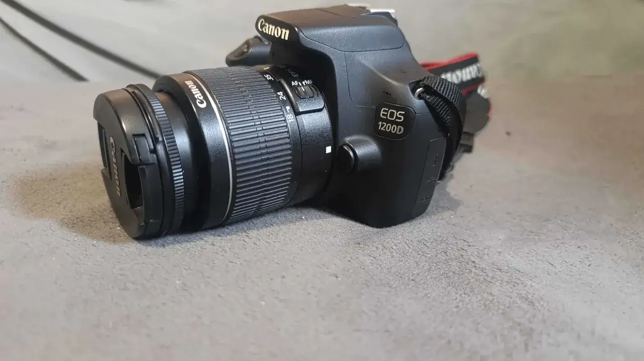 Czarny aparat Canon EOS 1200D z obiektywem leży na szarym tle.