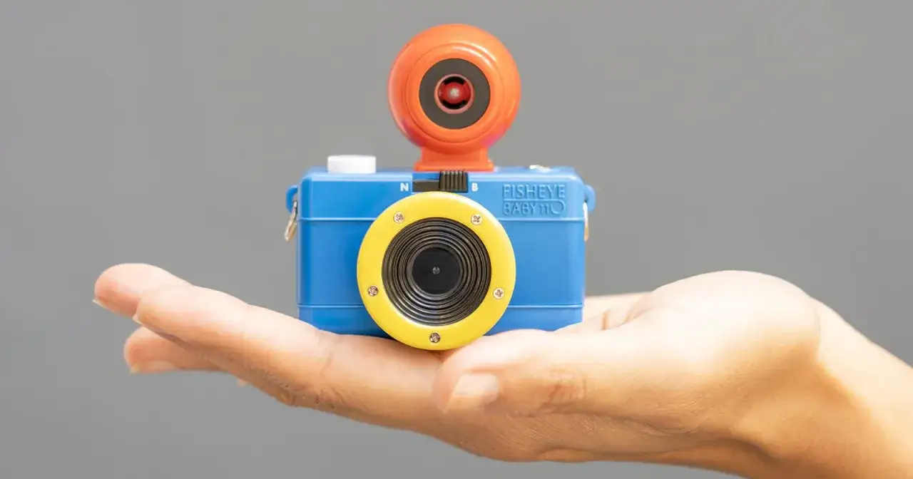 Niebieski aparat Fisheye Baby 110, idealny do lomografii, spoczywa na dłoni. Pomarańczowa nakładka na obiektyw dodaje mu uroku.