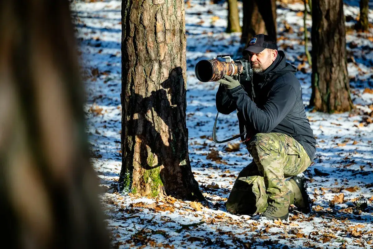 Fotograf przyrody w kamuflażu kuca przy drzewie, celując aparatem w zimowy las.