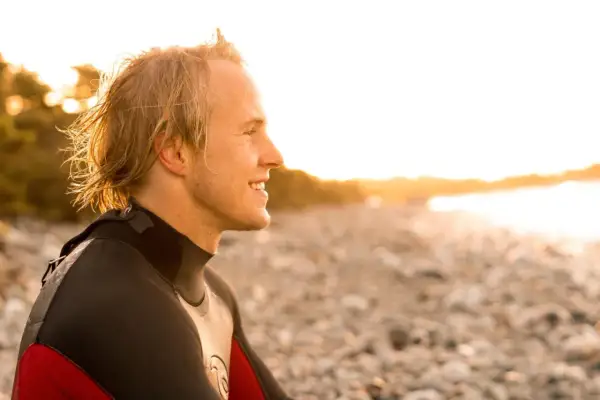 Uśmiechnięty surfer w piance, na planie średnim, patrzy w stronę zachodzącego słońca na kamienistej plaży.