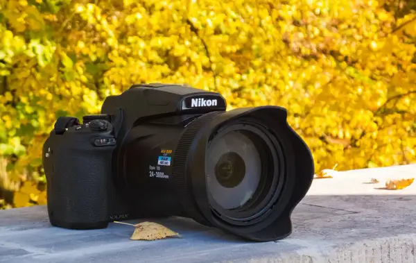 Czarny aparat Nikon P1000 z obiektywem na tle jesiennych, żółtych liści.