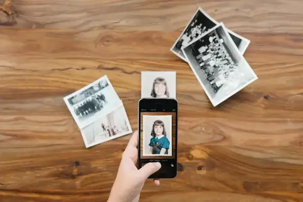 Skan ze zdjęcia: dłoń trzyma smartfon, który fotografuje zdjęcie dziewczynki. Wokół leżą stare fotografie.