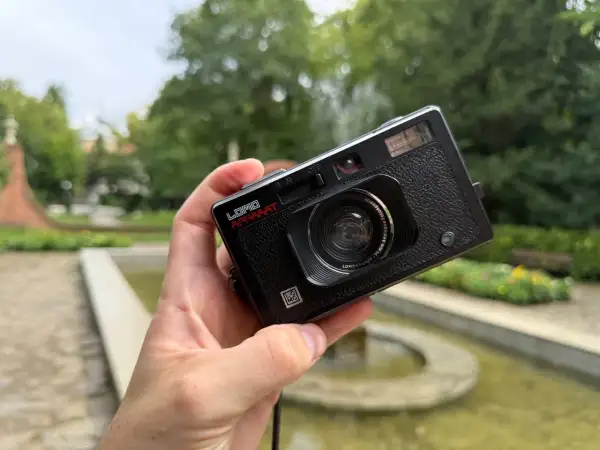 Czarna, analogowa lomografia LomoApparat z obiektywem 21mm. W dłoni, na tle zieleni i fontanny.
