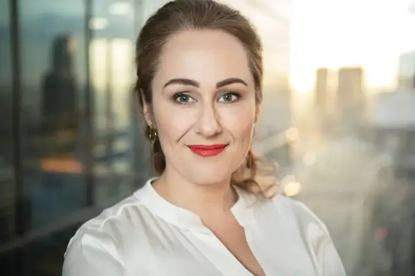 Portret fotografia: uśmiechnięta kobieta z burzą loków, w białej bluzce, z czerwonymi ustami, na tle rozmazanego miasta.