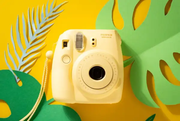 Żółty aparat Fujifilm Instax mini 8, z którego wychodzą zdjęcia, otoczony zielonymi liśćmi monstery i niebieskimi liśćmi paproci na żółtym tle.