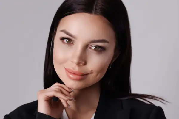 Kobieta w czarnej marynarce, z dłonią pod brodą, pokazuje, jak zrobić zdjęcie portretowe. Jej spojrzenie jest ciepłe, a usta lekko uchylone.