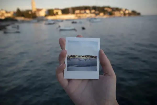 Dłoń trzyma zdjęcie jak z Instax z widokiem na zatokę z łodziami i miasteczkiem w tle.
